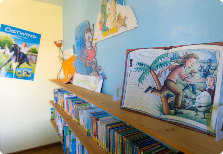 Holzregal voller Kinderb&uuml;cher; daneben ein aufgeklapptes Bilderbuch mit bunter Illustration an einer hellblauen Wand.
