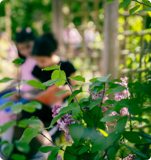 Fliederzweige im Vordergrund, unscharf im Hintergrund zwei Kinder beim Lesen im Schulgarten.