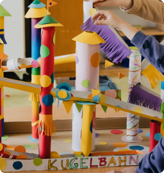 Bunte Kugelbahn aus bemalten Pappröhren, an der ein Kind gerade weiterbaut.