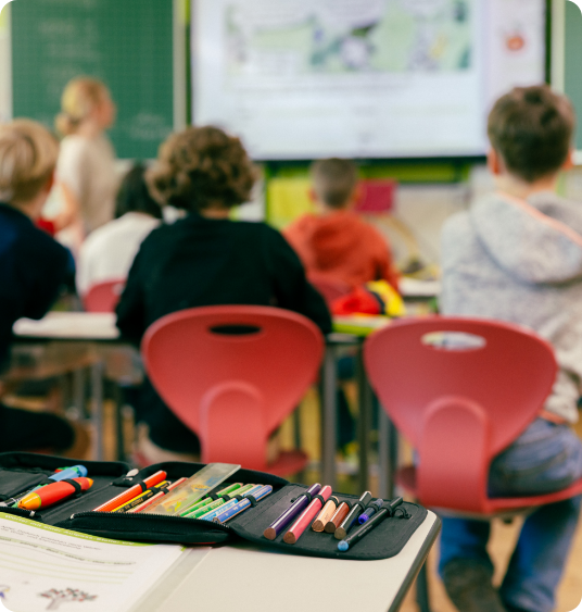 Schulkinder sitzen auf roten Stühlen und schauen zur Tafel; im Vordergrund liegt ein Etui mit Buntstiften.