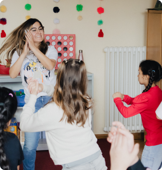 Lehrerin tanzt und klatscht fröhlich mit mehreren Kindern in einem Spielzimmer.