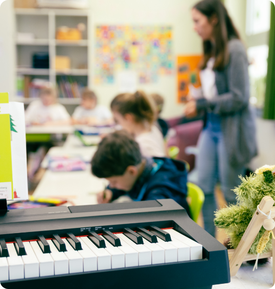 Elektrisches Keyboard im Klassenraum; Schüler arbeiten an Gruppentischen, Lehrerin betreut.